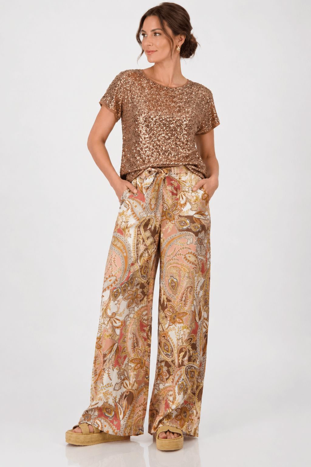 Monari modeling a short-sleeve crewneck top covered in shimmering rose gold sequins, paired with brown trousers, against a plain light gray studio background.