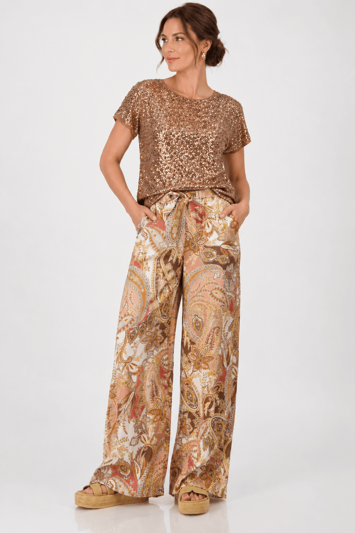 Monari modeling a short-sleeve crewneck top covered in shimmering rose gold sequins, paired with brown trousers, against a plain light gray studio background.