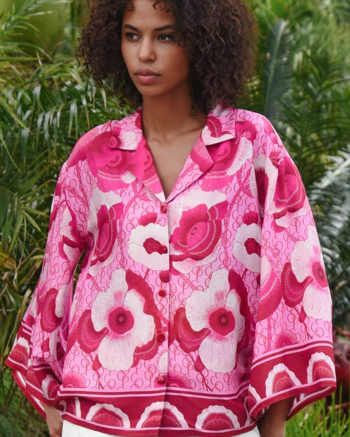 Trelise Cooper model wearing a vibrant pink and red floral print button-up shirt with white pants, standing outdoors in front of a lush green tropical background.