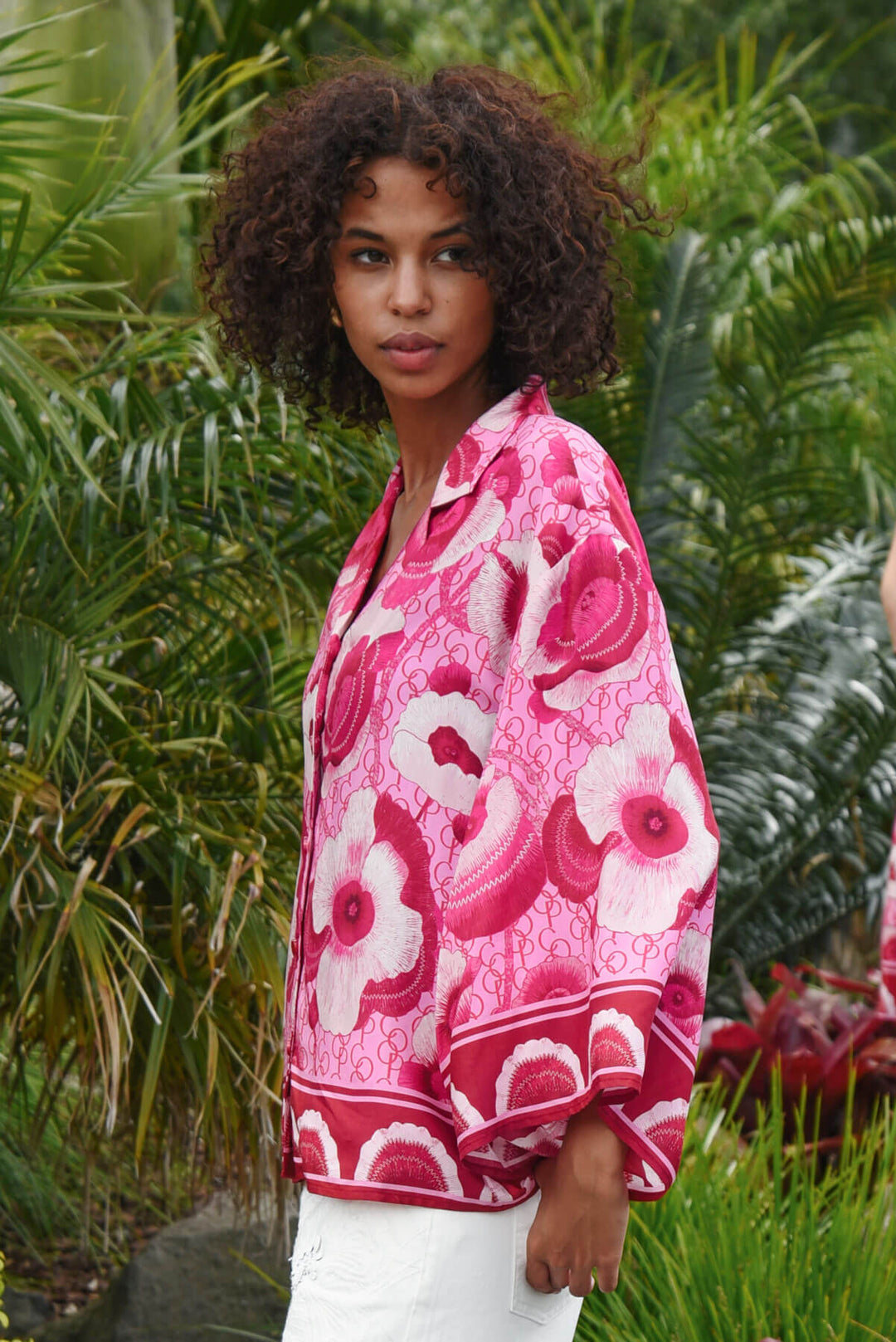 Trelise Cooper model wearing a vibrant pink and red floral print button-up shirt with white pants, standing outdoors in front of a lush green tropical background.