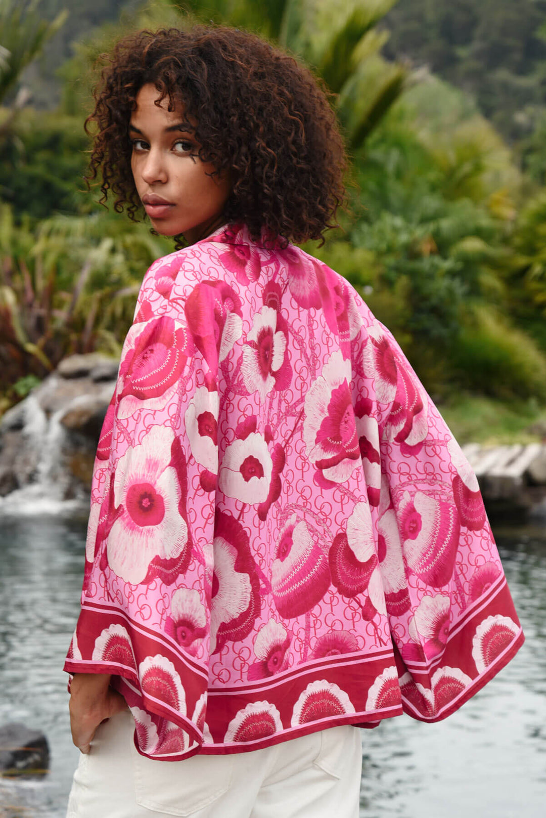 Trelise Cooper model wearing a vibrant pink and red floral print button-up shirt with white pants, standing outdoors in front of a lush green tropical background. Back view. 