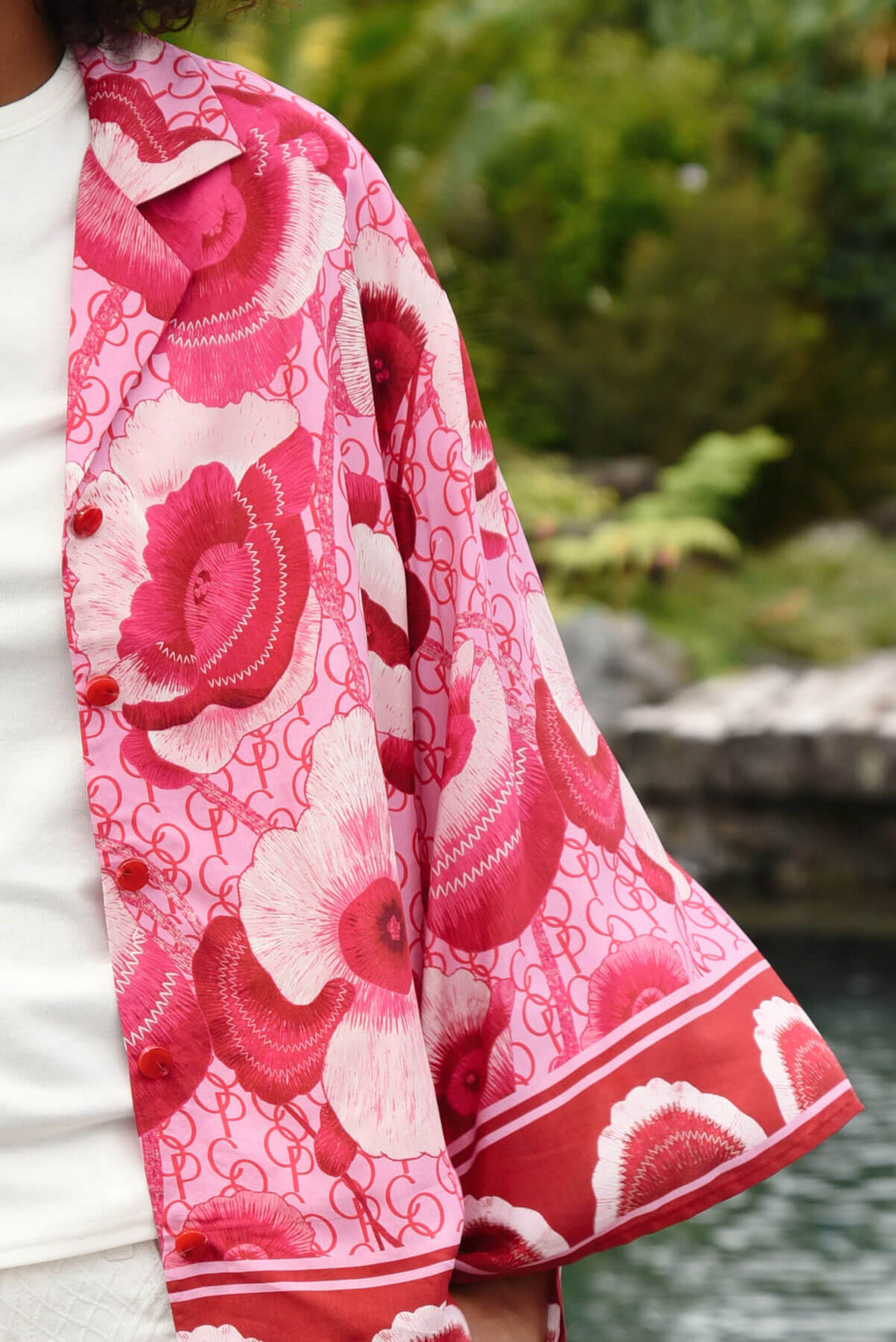 Trelise Cooper model wearing a vibrant pink and red floral print button-up shirt with white pants, standing outdoors in front of a lush green tropical background. Sleeve. 