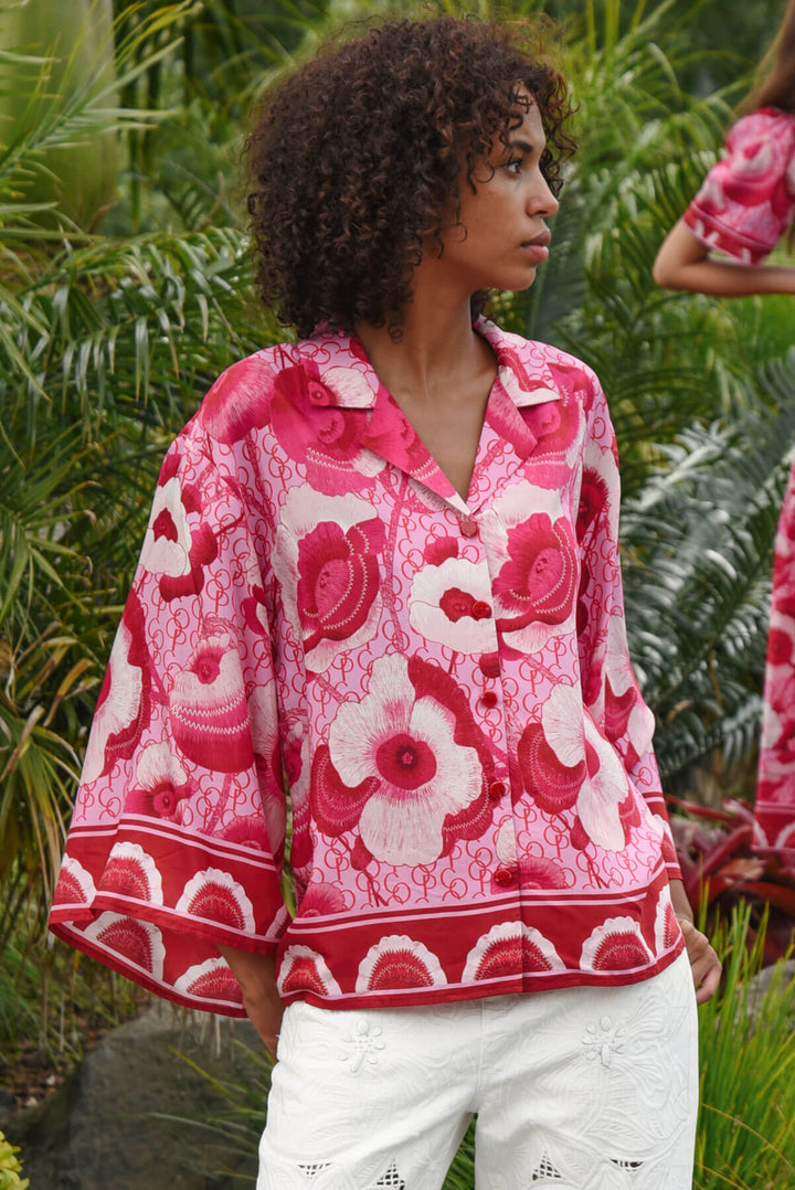 Trelise Cooper model wearing a vibrant pink and red floral print button-up shirt with white pants, standing outdoors in front of a lush green tropical background.