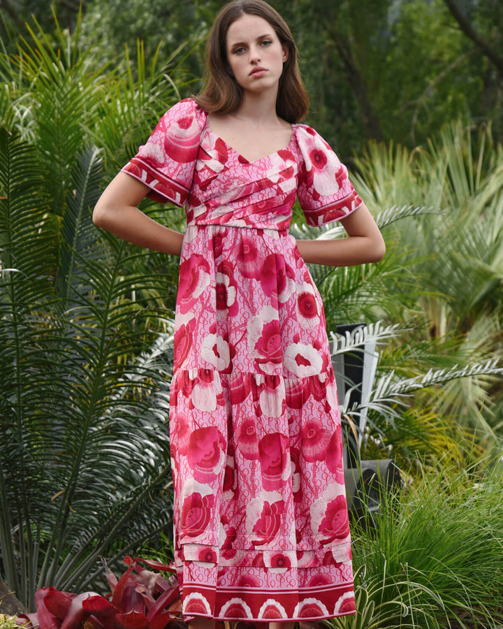Trelise Cooper Model  stands barefoot outdoors among tropical green plants, wearing a pink floral midi dress with short flutter sleeves and a gathered waist.