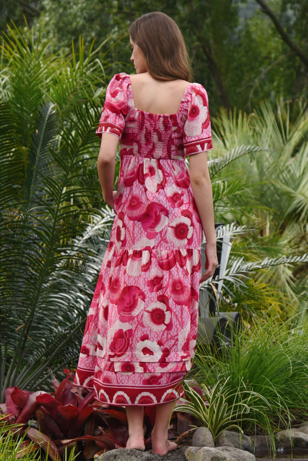 Trelise Cooper Model  stands barefoot outdoors among tropical green plants, wearing a pink floral midi dress with short flutter sleeves and a gathered waist. Back view. 