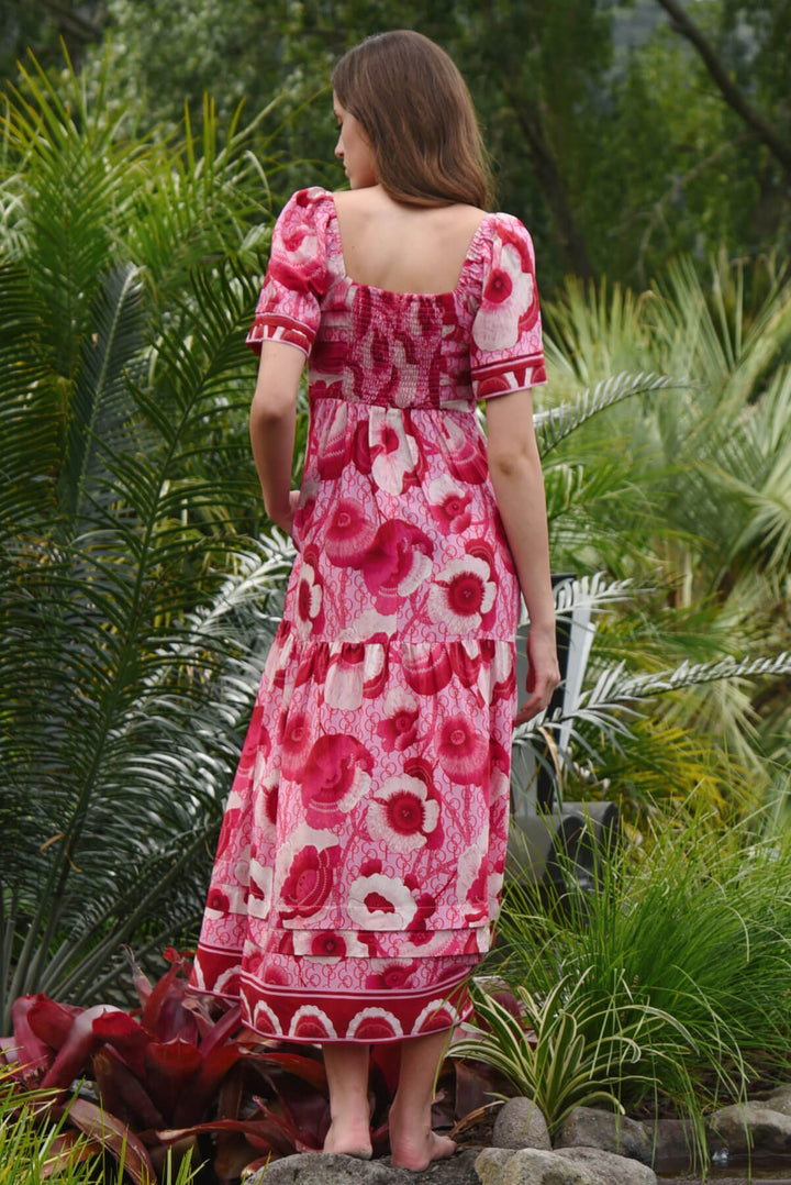 Trelise Cooper Model  stands barefoot outdoors among tropical green plants, wearing a pink floral midi dress with short flutter sleeves and a gathered waist. Back view. 
