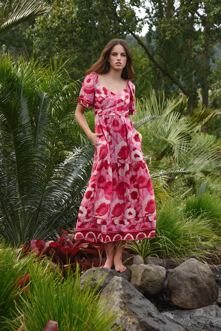 Trelise Cooper Model  stands barefoot outdoors among tropical green plants, wearing a pink floral midi dress with short flutter sleeves and a gathered waist.