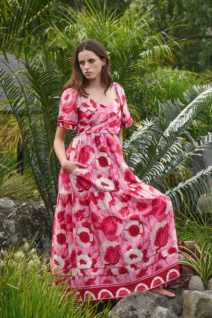 Trelise Cooper Model  stands barefoot outdoors among tropical green plants, wearing a pink floral midi dress with short flutter sleeves and a gathered waist.