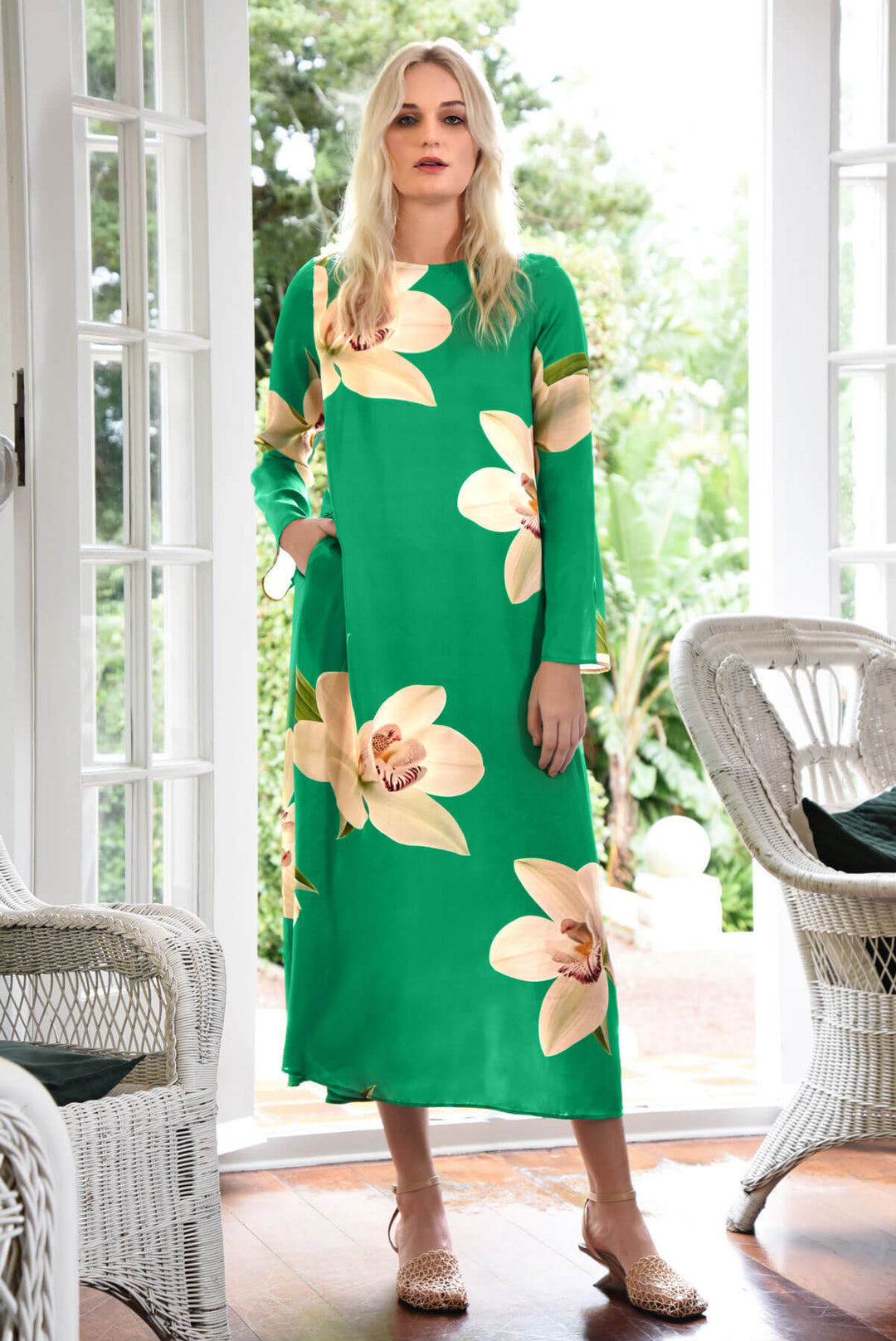 Trelise Cooper  modeling a green long-sleeve midi dress with large beige floral prints, standing in a bright room with white wicker chairs and glass doors.