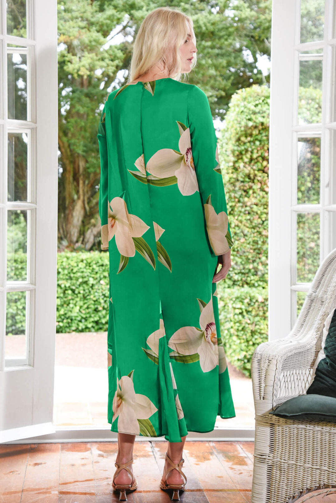 Trelise Cooper  modeling a green long-sleeve midi dress with large beige floral prints, standing in a bright room with white wicker chairs and glass doors. back view