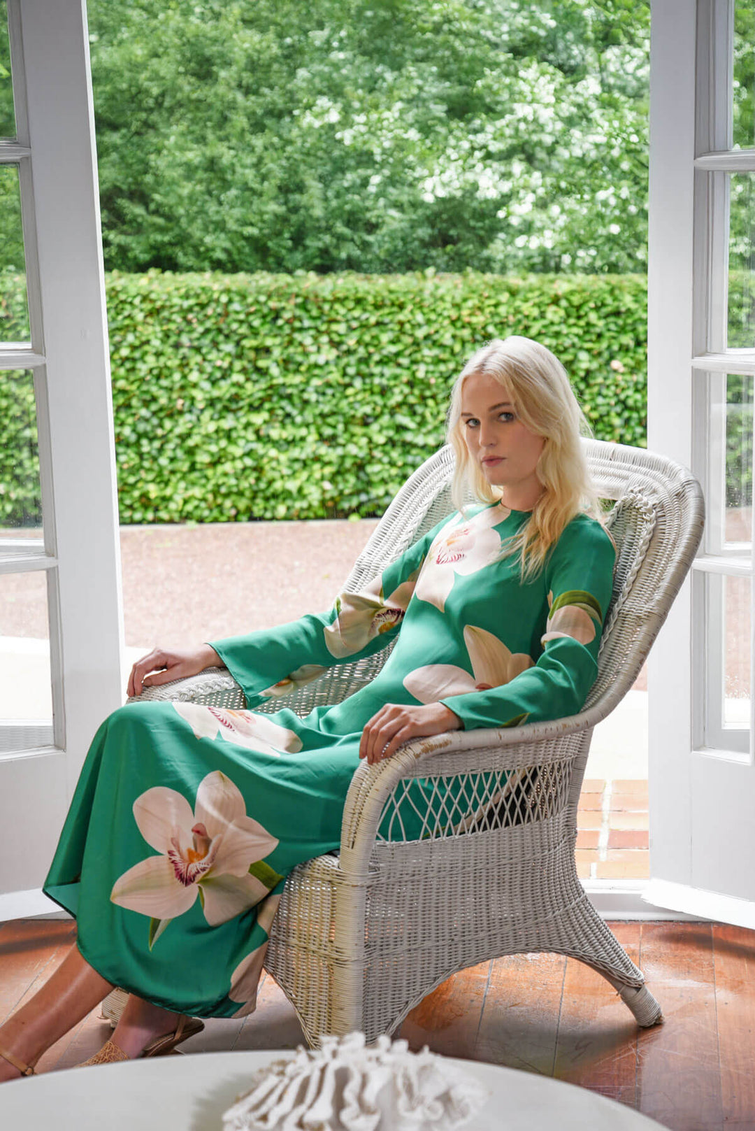 Trelise Cooper  modeling a green long-sleeve midi dress with large beige floral prints, standing in a bright room with white wicker chairs and glass doors. Sitting down