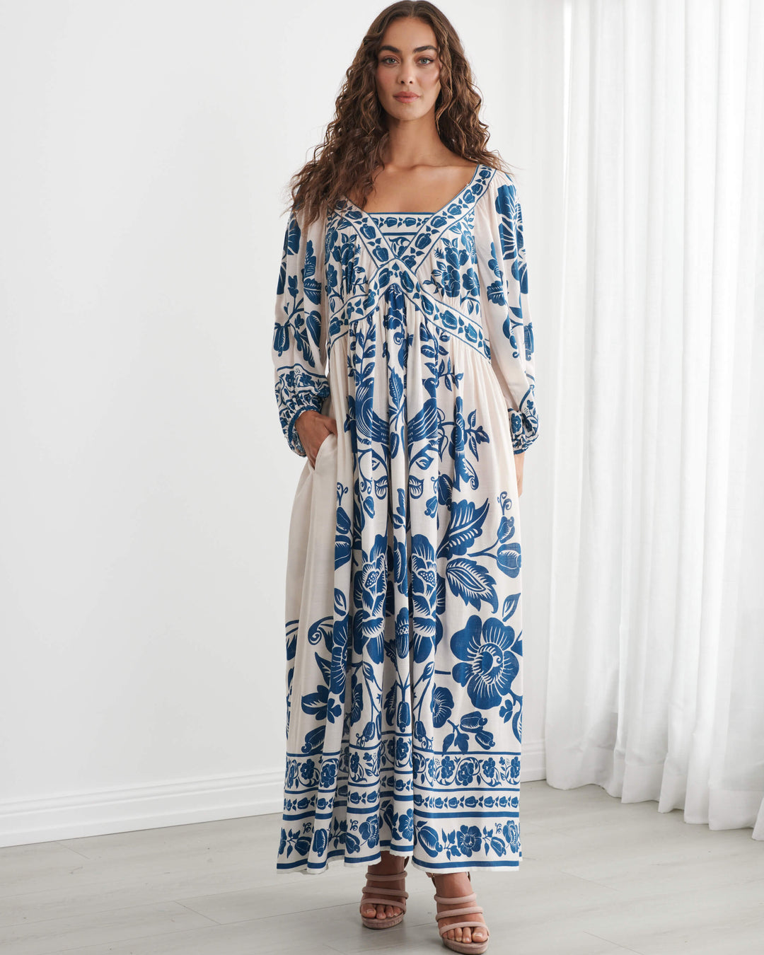 Caju model wearing a long-sleeved bohemian maxi dress with a blue and cream floral pattern, standing in a bright room with white walls and sheer curtains. On a white background
