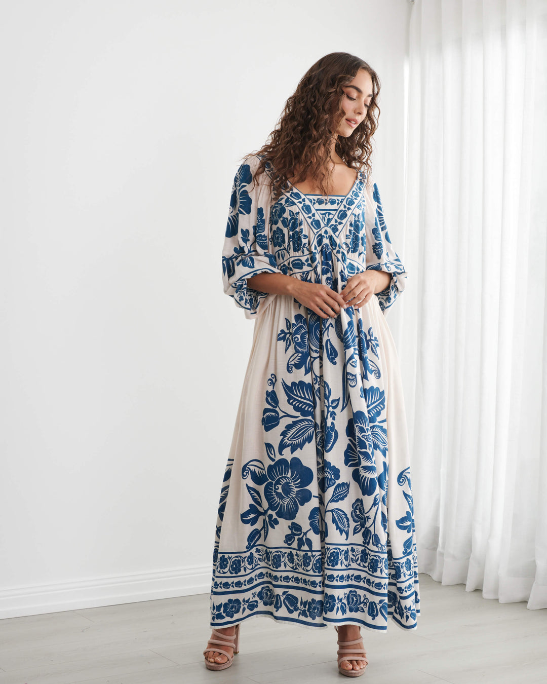 Caju model wearing a long-sleeved bohemian maxi dress with a blue and cream floral pattern, standing in a bright room with white walls and sheer curtains. On a white background