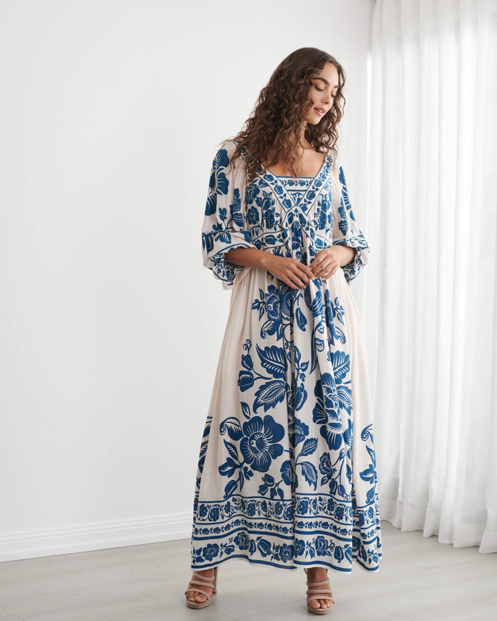 Caju model wearing a long-sleeved bohemian maxi dress with a blue and cream floral pattern, standing in a bright room with white walls and sheer curtains. On a white background