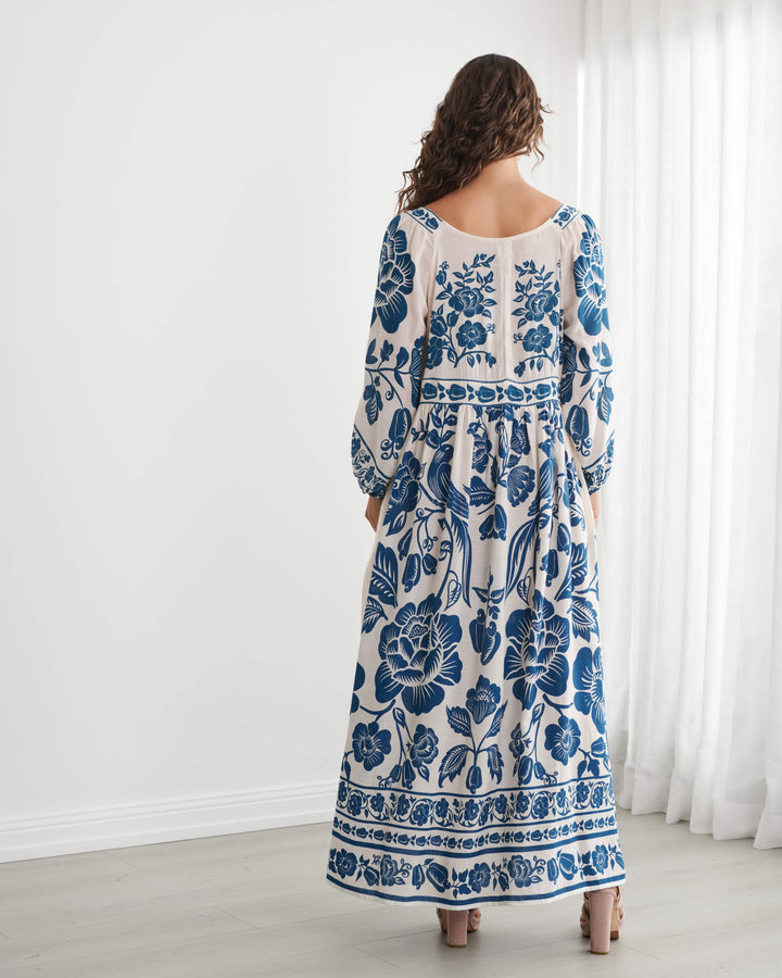 Caju model wearing a long-sleeved bohemian maxi dress with a blue and cream floral pattern, standing in a bright room with white walls and sheer curtains. On a white background