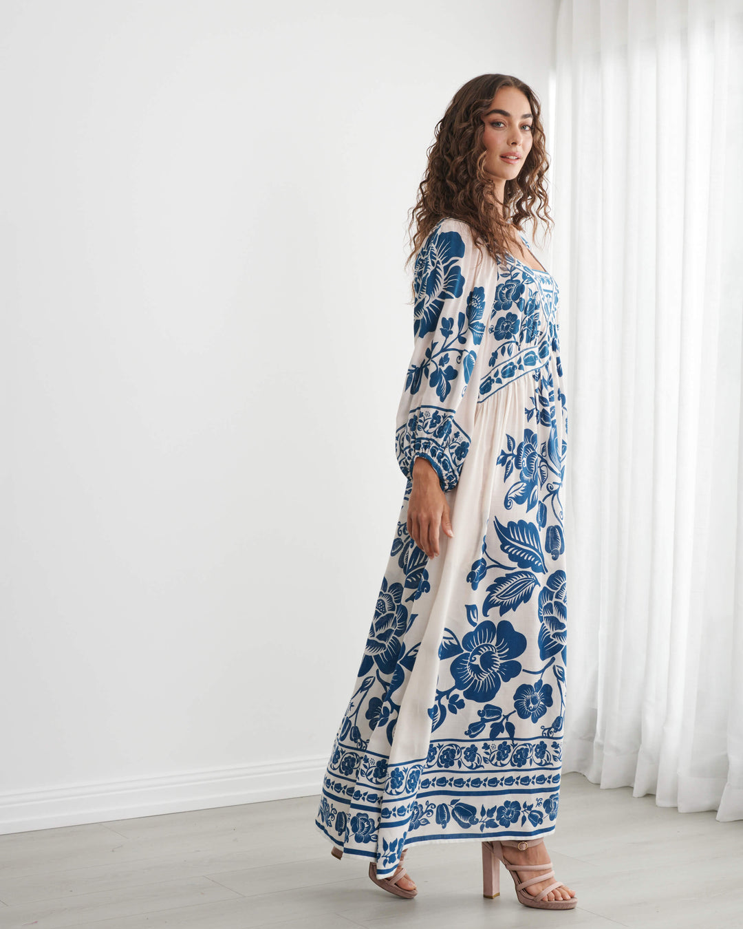 Caju model wearing a long-sleeved bohemian maxi dress with a blue and cream floral pattern, standing in a bright room with white walls and sheer curtains. On a white background