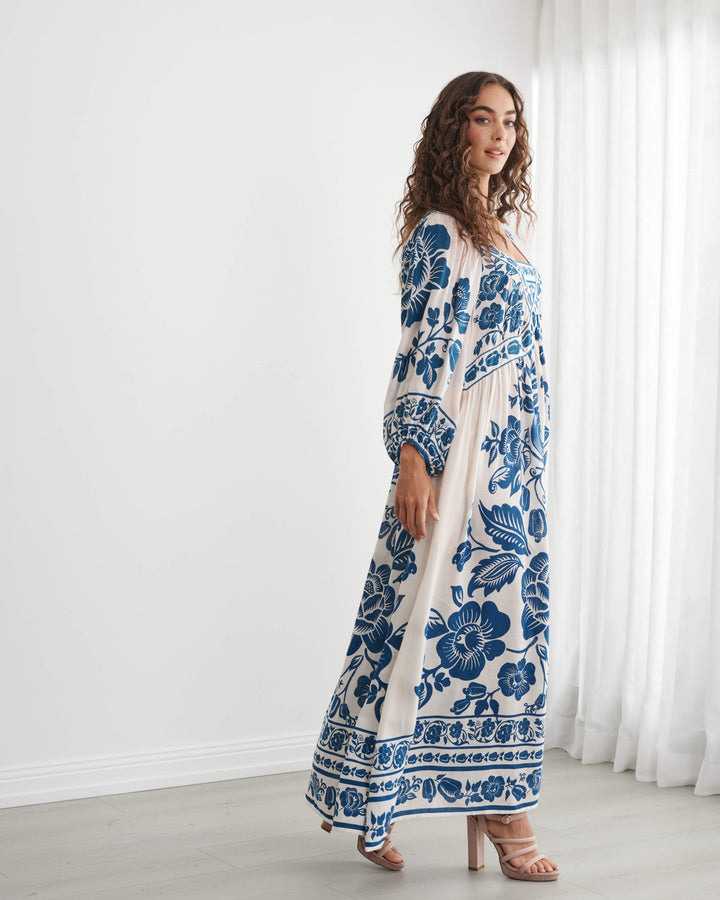 Caju model wearing a long-sleeved bohemian maxi dress with a blue and cream floral pattern, standing in a bright room with white walls and sheer curtains. On a white background