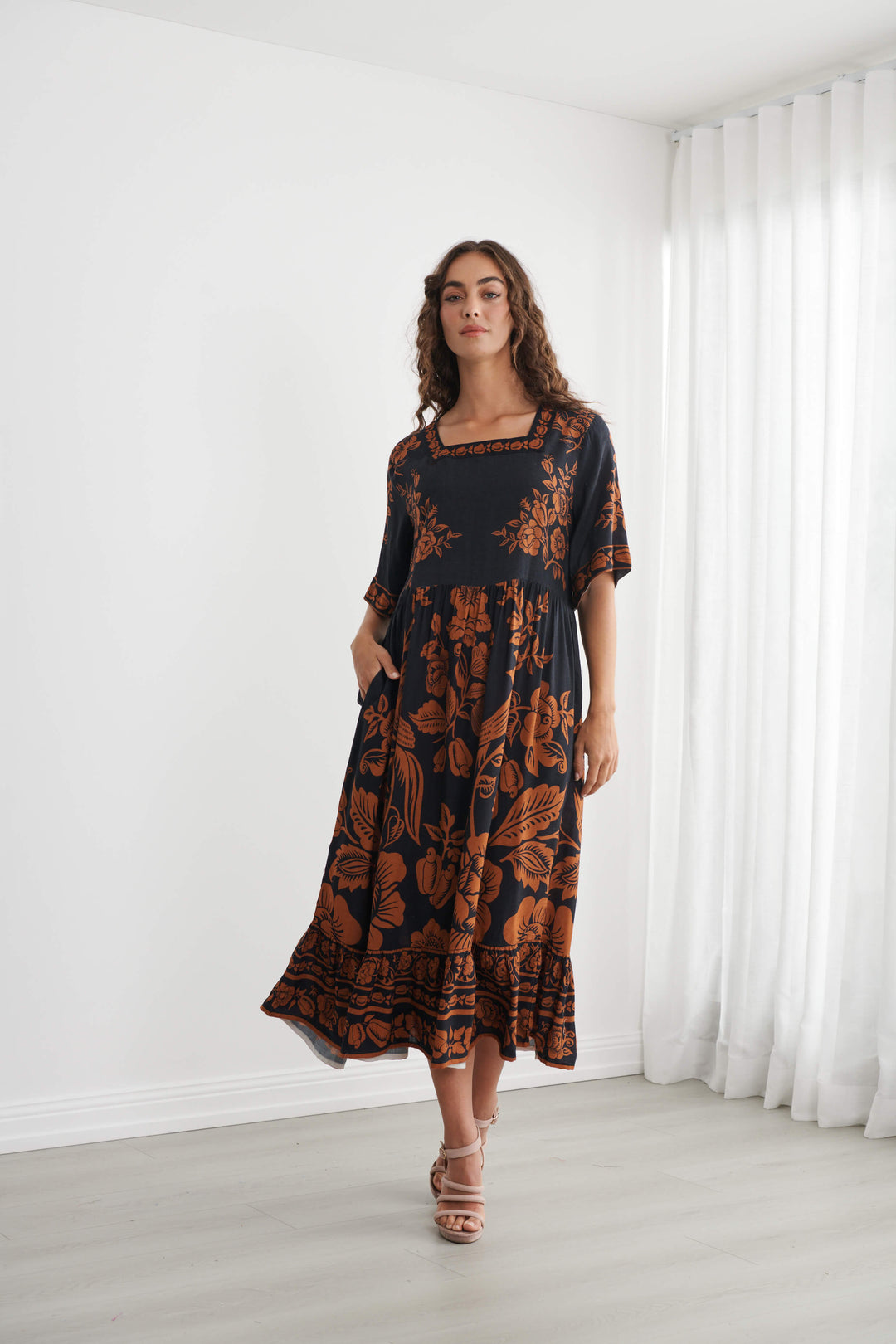 Caju A woman modeling a black midi dress with a rust-orange floral and botanical pattern, standing in a bright room with white walls and sheer white curtains.