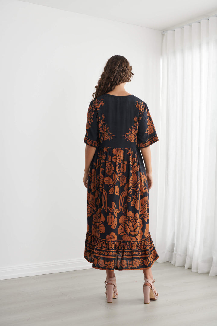 Caju A woman modeling a black midi dress with a rust-orange floral and botanical pattern, standing in a bright room with white walls and sheer white curtains. Back view 