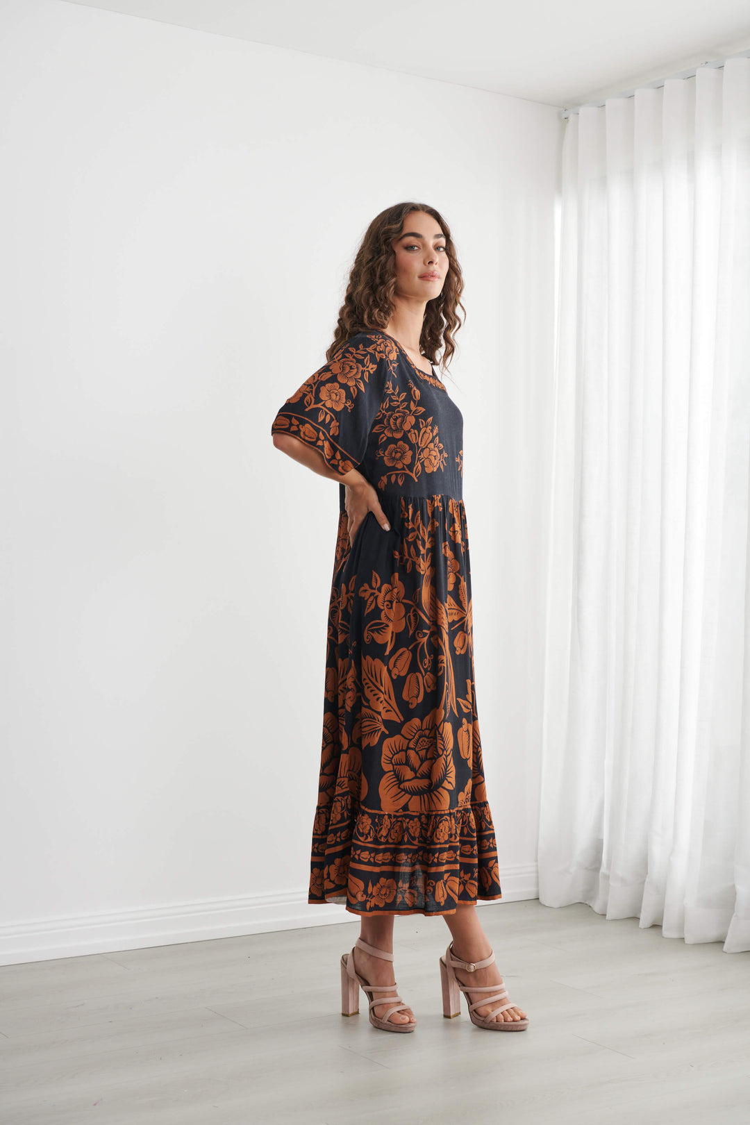 Caju A woman modeling a black midi dress with a rust-orange floral and botanical pattern, standing in a bright room with white walls and sheer white curtains.