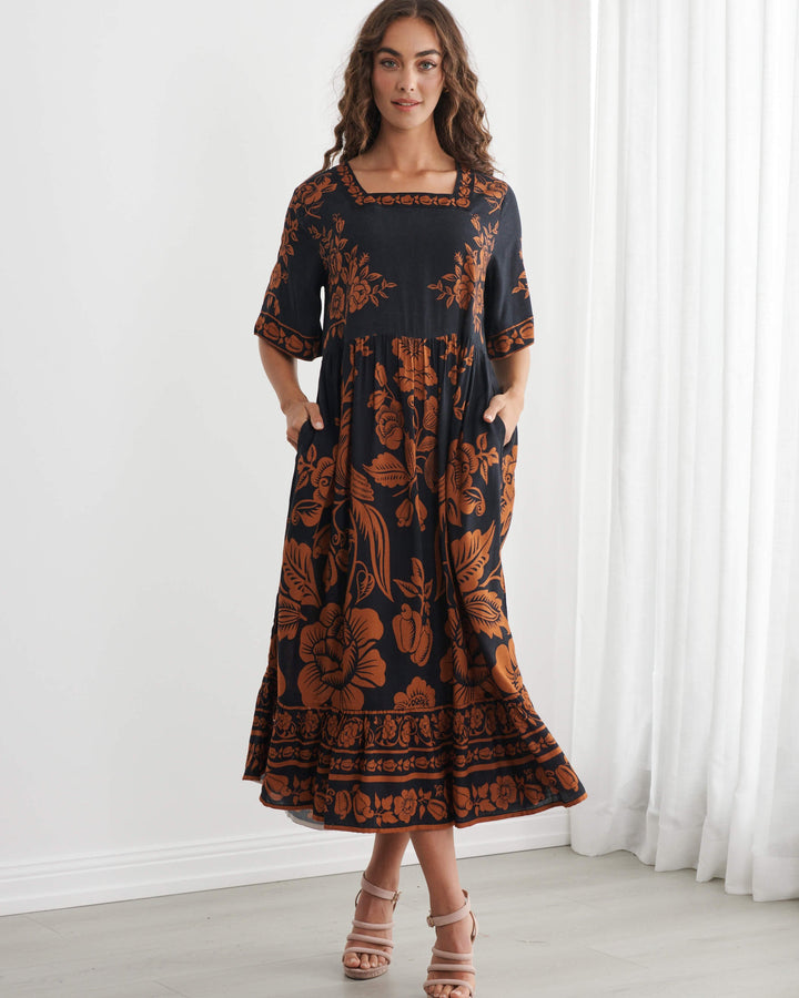 Caju A woman modeling a black midi dress with a rust-orange floral and botanical pattern, standing in a bright room with white walls and sheer white curtains.