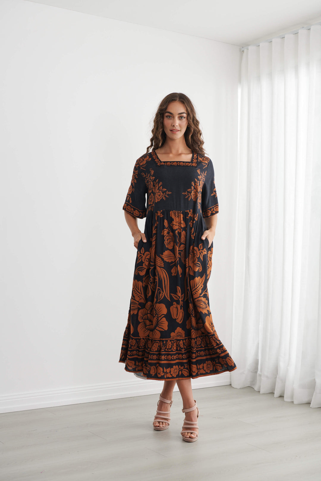 Caju A woman modeling a black midi dress with a rust-orange floral and botanical pattern, standing in a bright room with white walls and sheer white curtains.