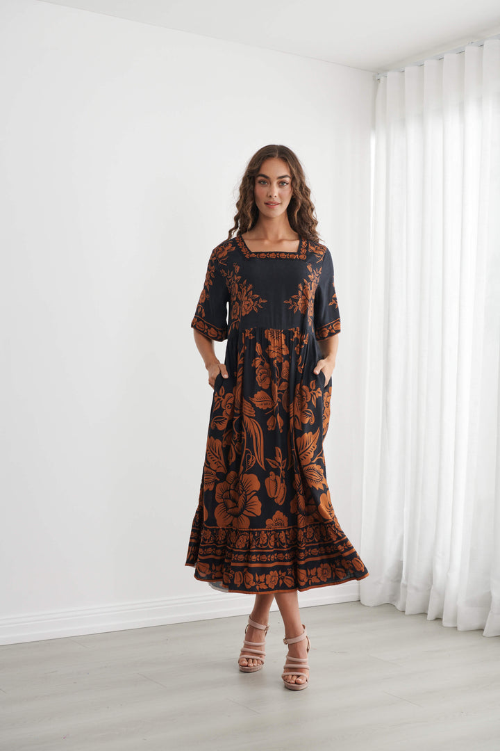 Caju A woman modeling a black midi dress with a rust-orange floral and botanical pattern, standing in a bright room with white walls and sheer white curtains.