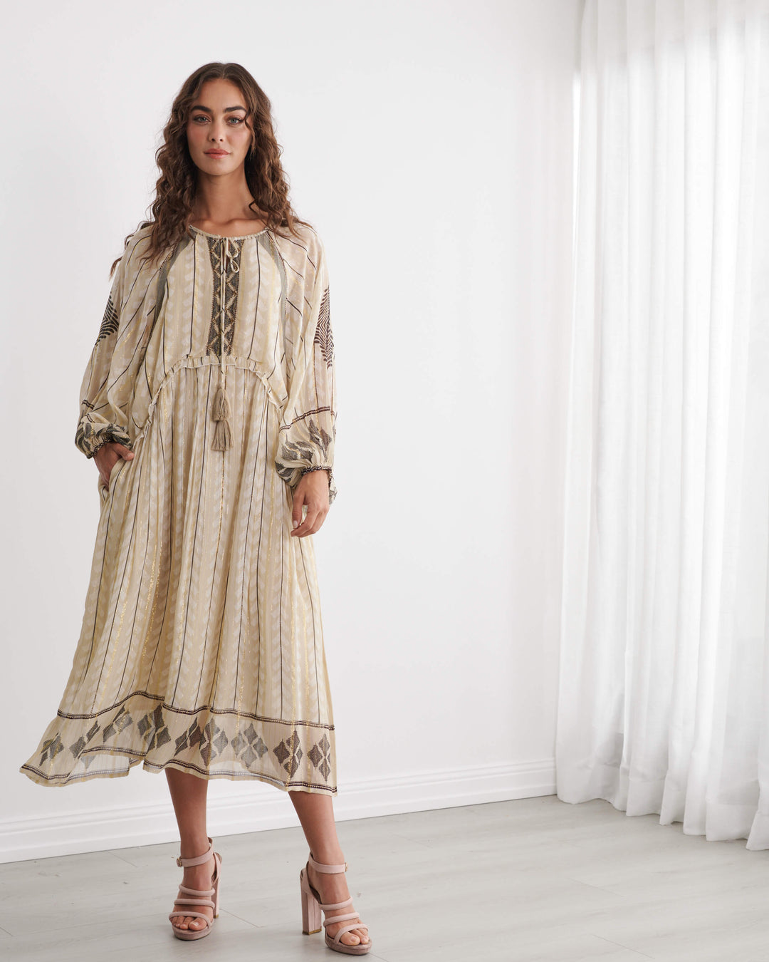 Caju woman modeling a long beige bohemian maxi dress with brown geometric patterns and lace-up detailing, standing in a bright white room with sheer curtains.