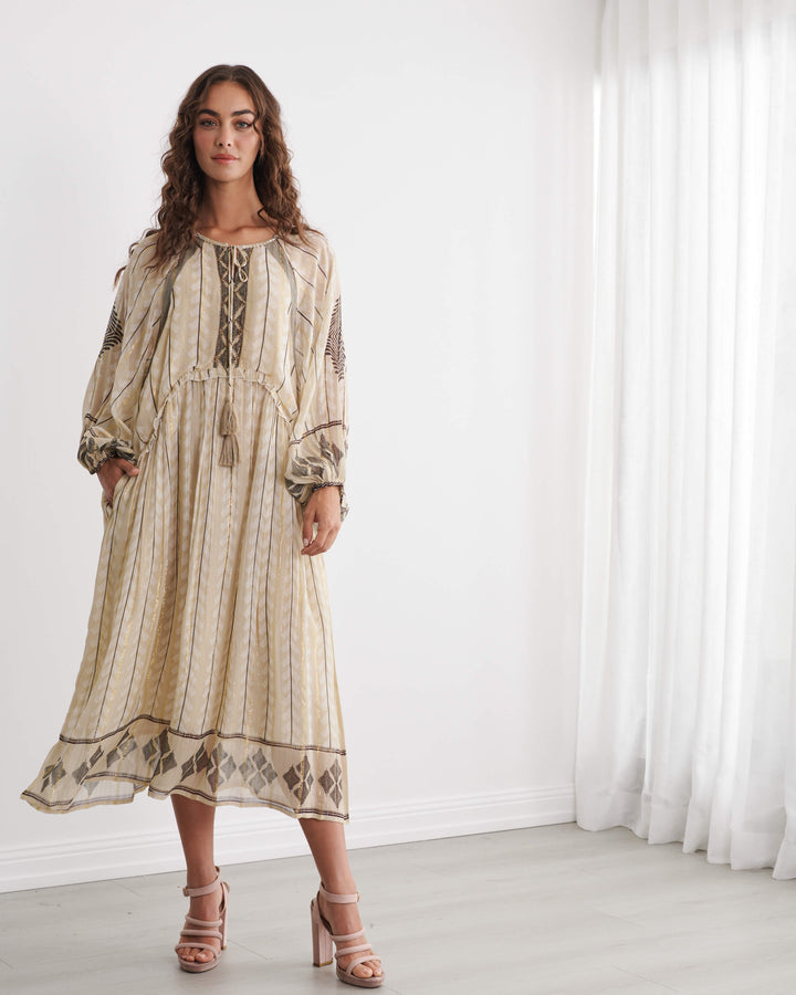Caju woman modeling a long beige bohemian maxi dress with brown geometric patterns and lace-up detailing, standing in a bright white room with sheer curtains.