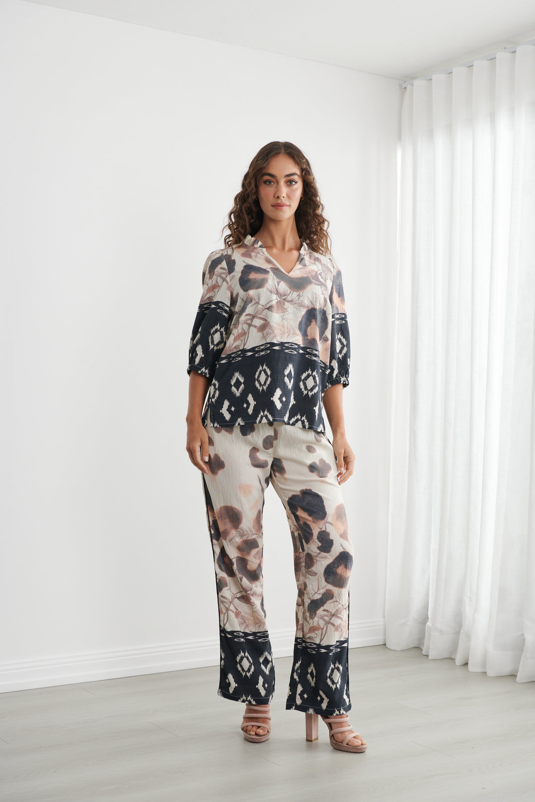 Caju models a matching two-piece lounge set featuring a beige, black, and brown animal print with black geometric lace trim, set against a bright white room with sheer curtains.