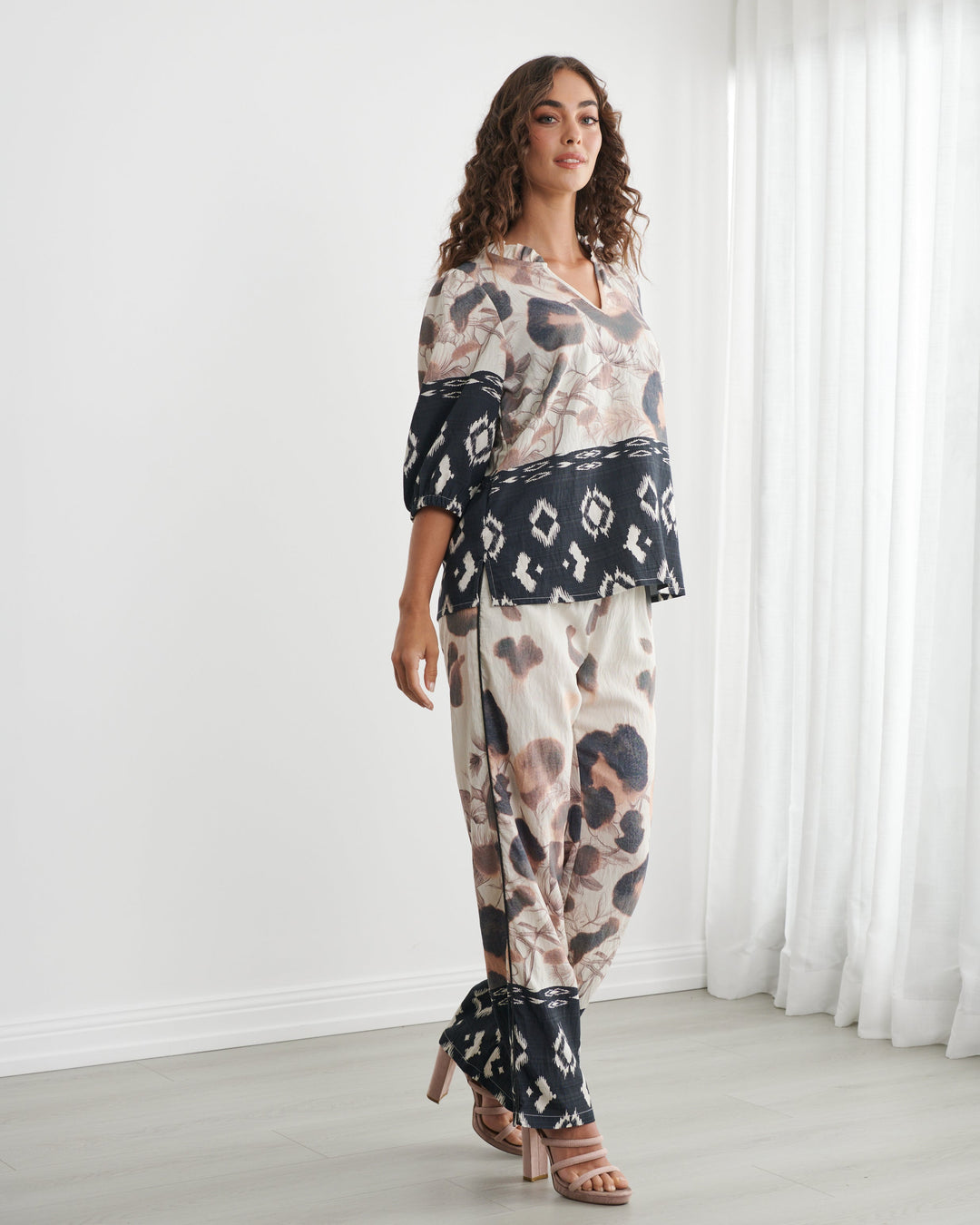 Caju models a matching two-piece lounge set featuring a beige, black, and brown animal print with black geometric lace trim, set against a bright white room with sheer curtains.