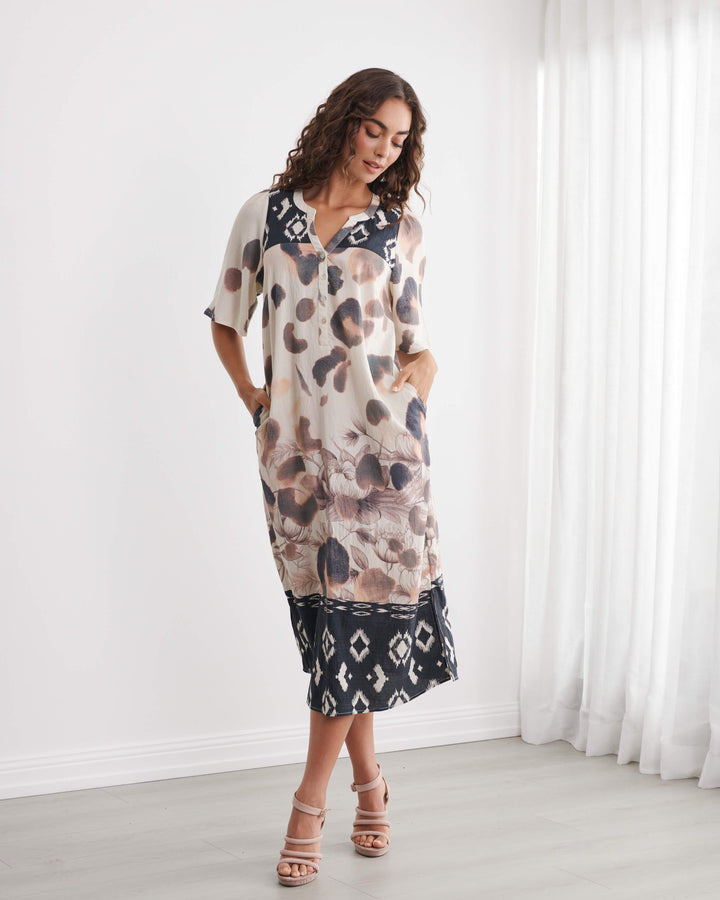 Caju models a short-sleeve midi dress with an abstract animal print in beige, brown, and black, featuring a contrasting black geometric pattern hem and collar, set against a bright white room background.