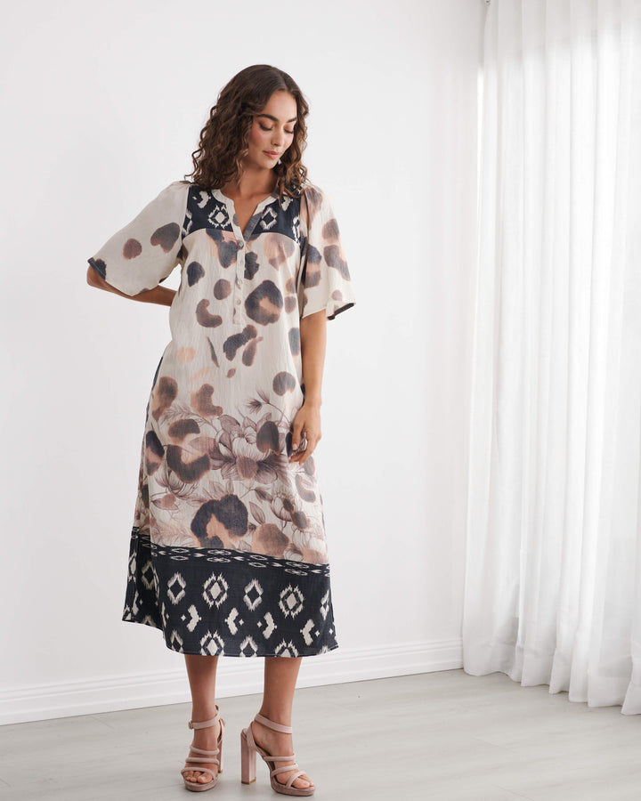 Caju models a short-sleeve midi dress with an abstract animal print in beige, brown, and black, featuring a contrasting black geometric pattern hem and collar, set against a bright white room background.