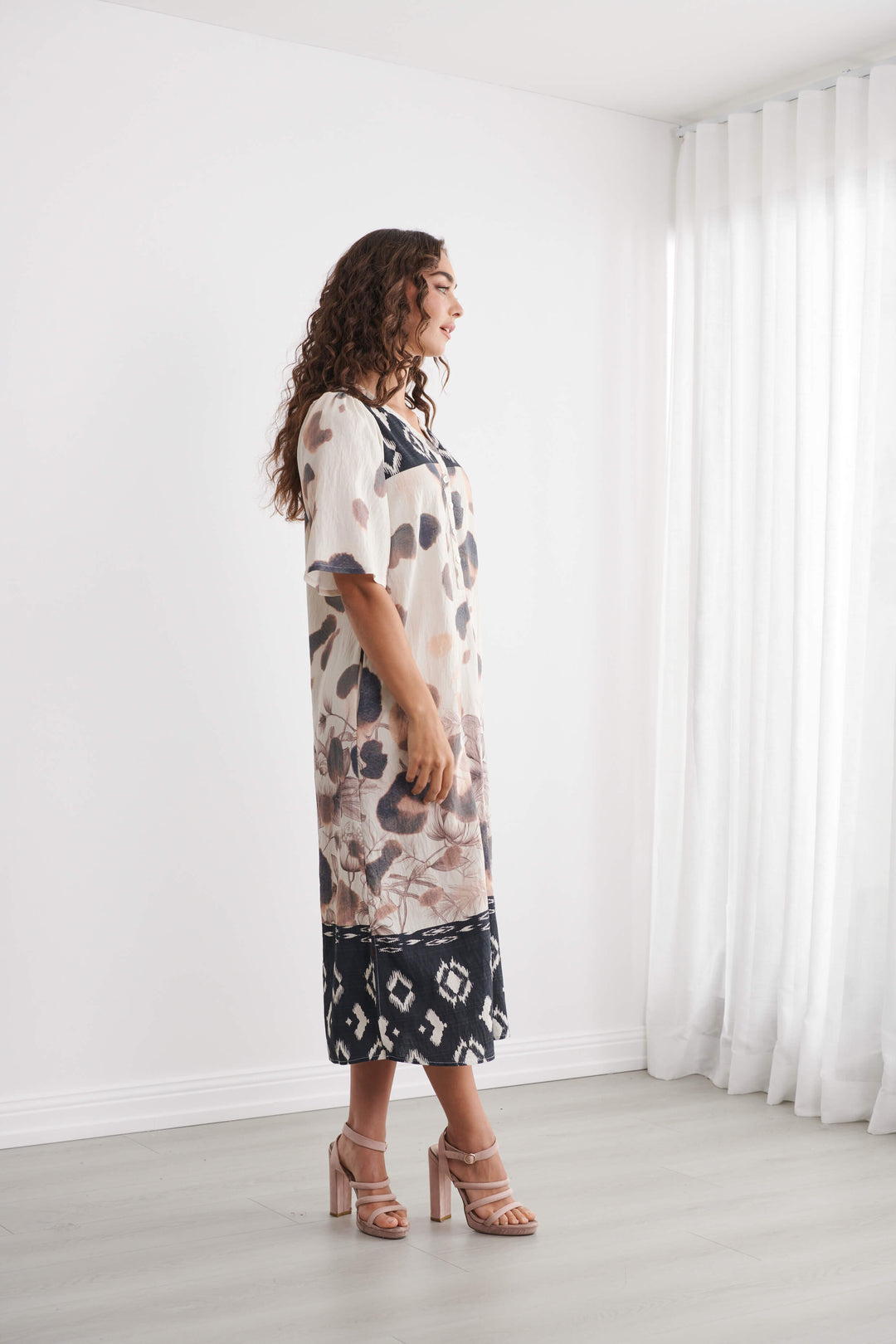 Caju models a short-sleeve midi dress with an abstract animal print in beige, brown, and black, featuring a contrasting black geometric pattern hem and collar, set against a bright white room background.