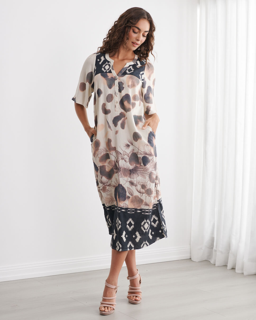 Caju models a short-sleeve midi dress with an abstract animal print in beige, brown, and black, featuring a contrasting black geometric pattern hem and collar, set against a bright white room background.