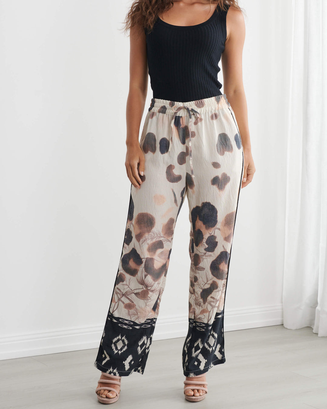 Caju modeling wide-leg pants with a beige, brown, and black abstract pattern, paired with a black tank top and neutral sandals, standing in a bright room with white walls and sheer curtains.