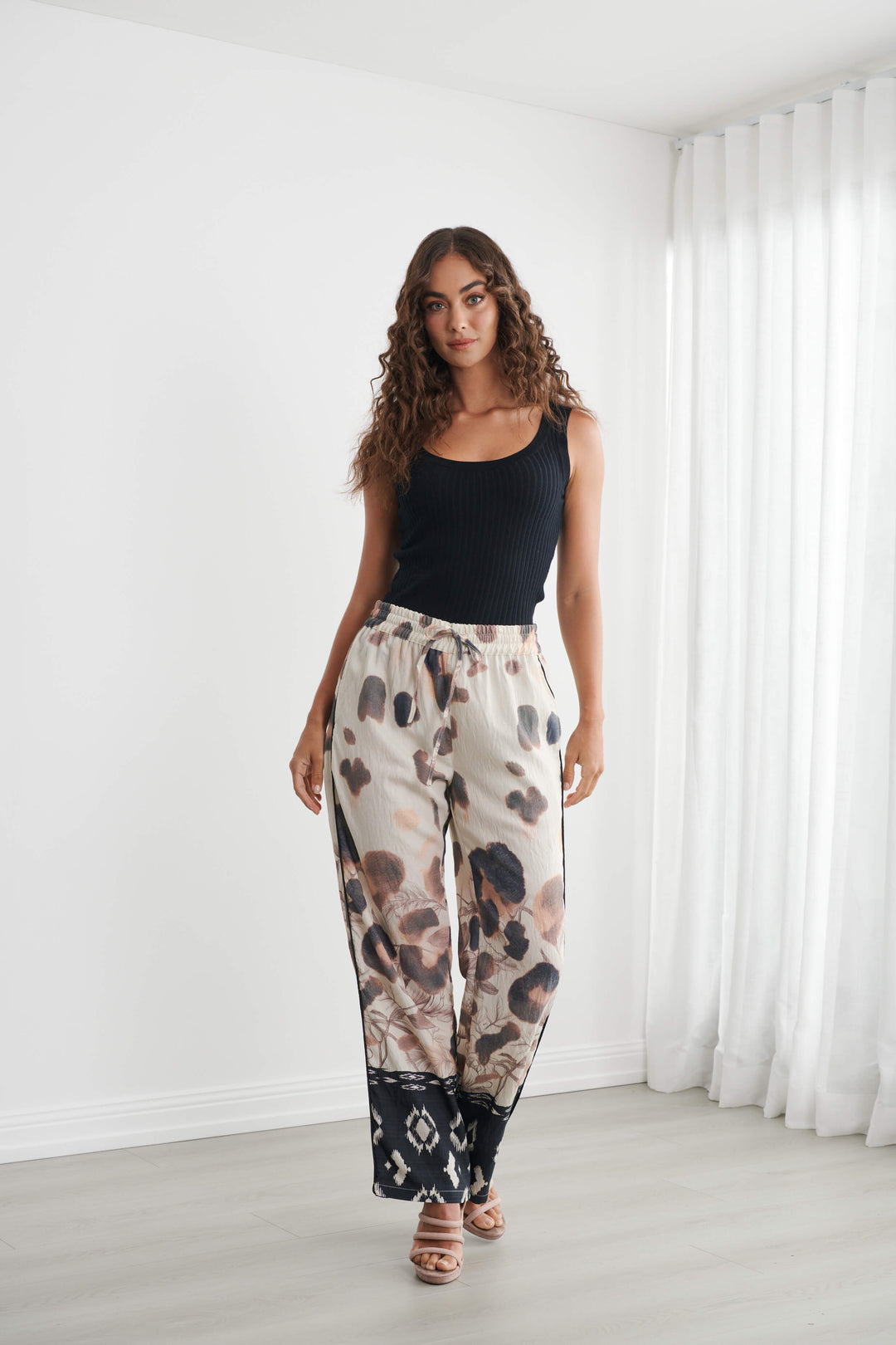 Caju modeling wide-leg pants with a beige, brown, and black abstract pattern, paired with a black tank top and neutral sandals, standing in a bright room with white walls and sheer curtains.