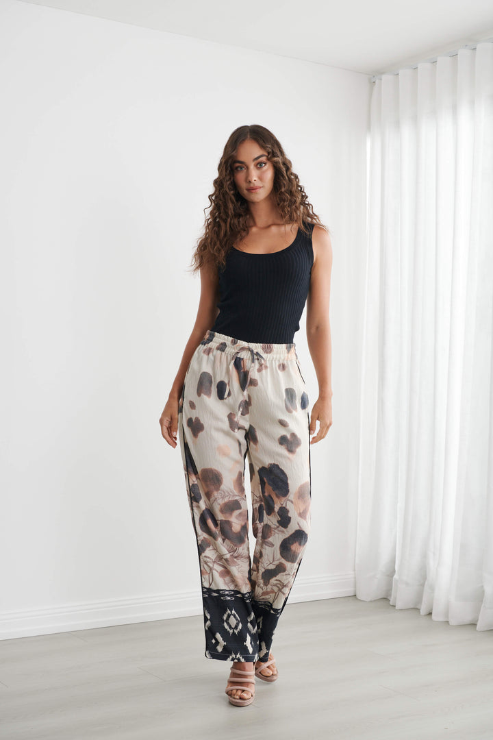 Caju modeling wide-leg pants with a beige, brown, and black abstract pattern, paired with a black tank top and neutral sandals, standing in a bright room with white walls and sheer curtains.