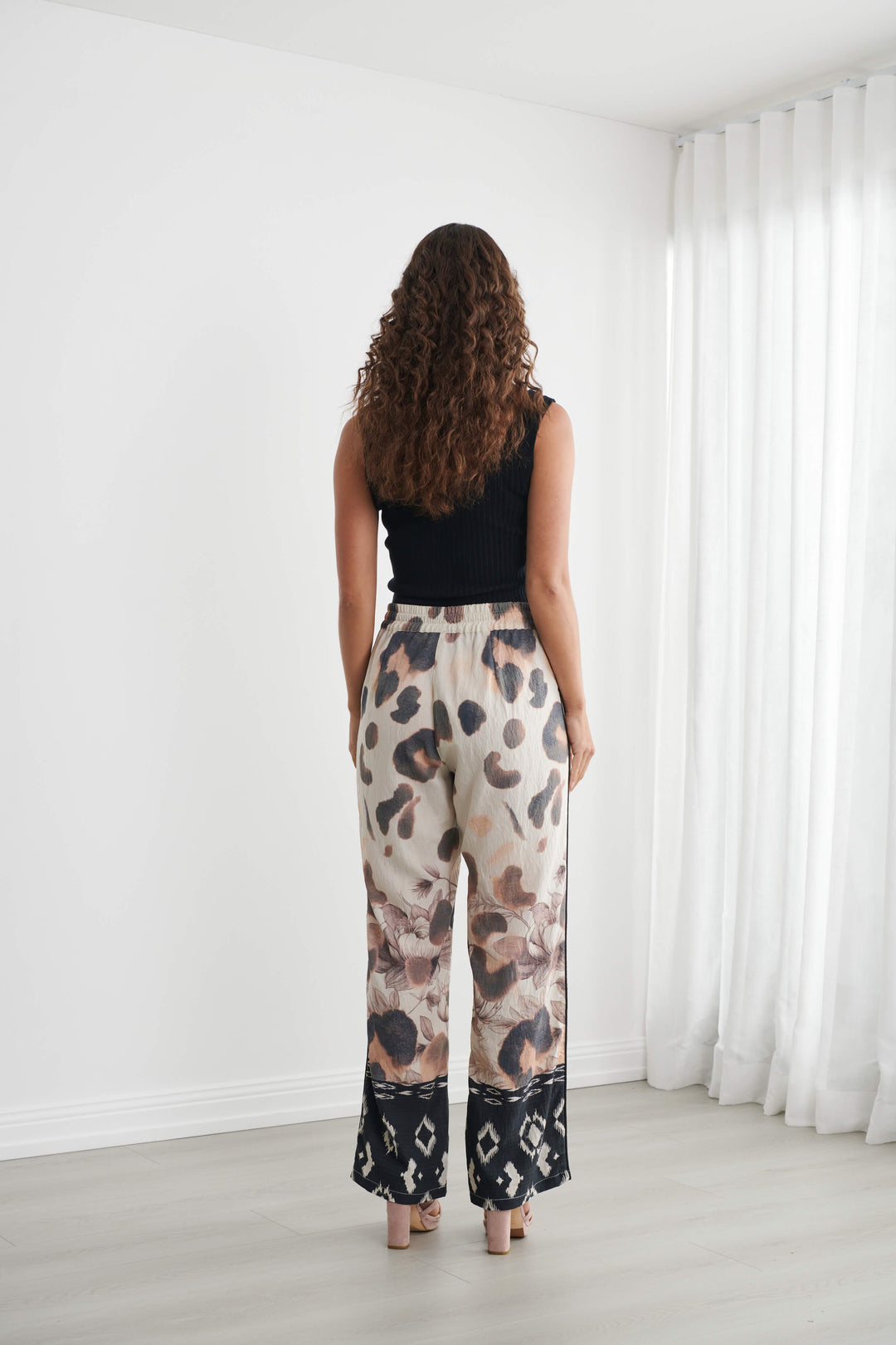 Caju modeling wide-leg pants with a beige, brown, and black abstract pattern, paired with a black tank top and neutral sandals, standing in a bright room with white walls and sheer curtains.
Back view 