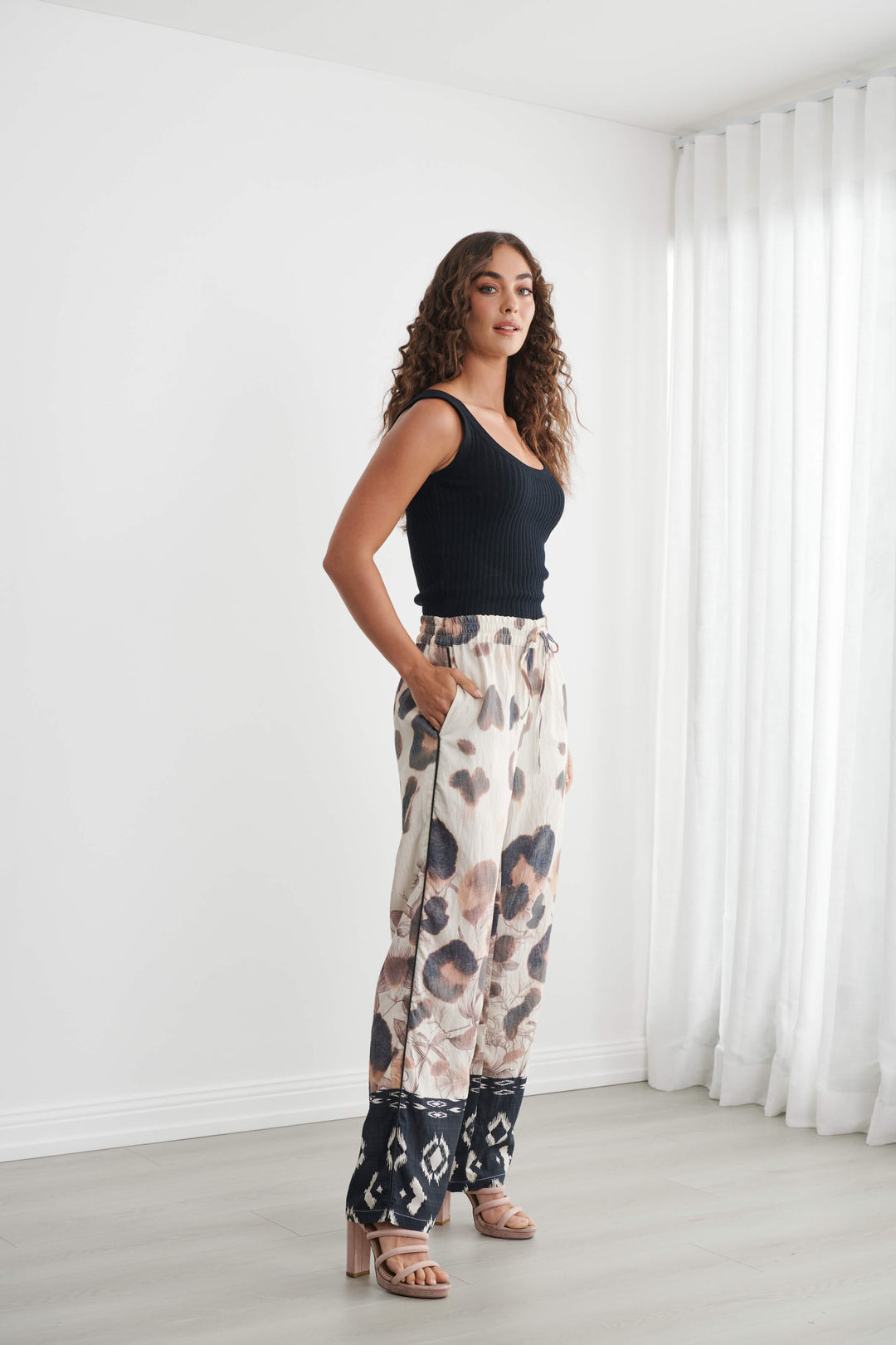 Caju modeling wide-leg pants with a beige, brown, and black abstract pattern, paired with a black tank top and neutral sandals, standing in a bright room with white walls and sheer curtains.