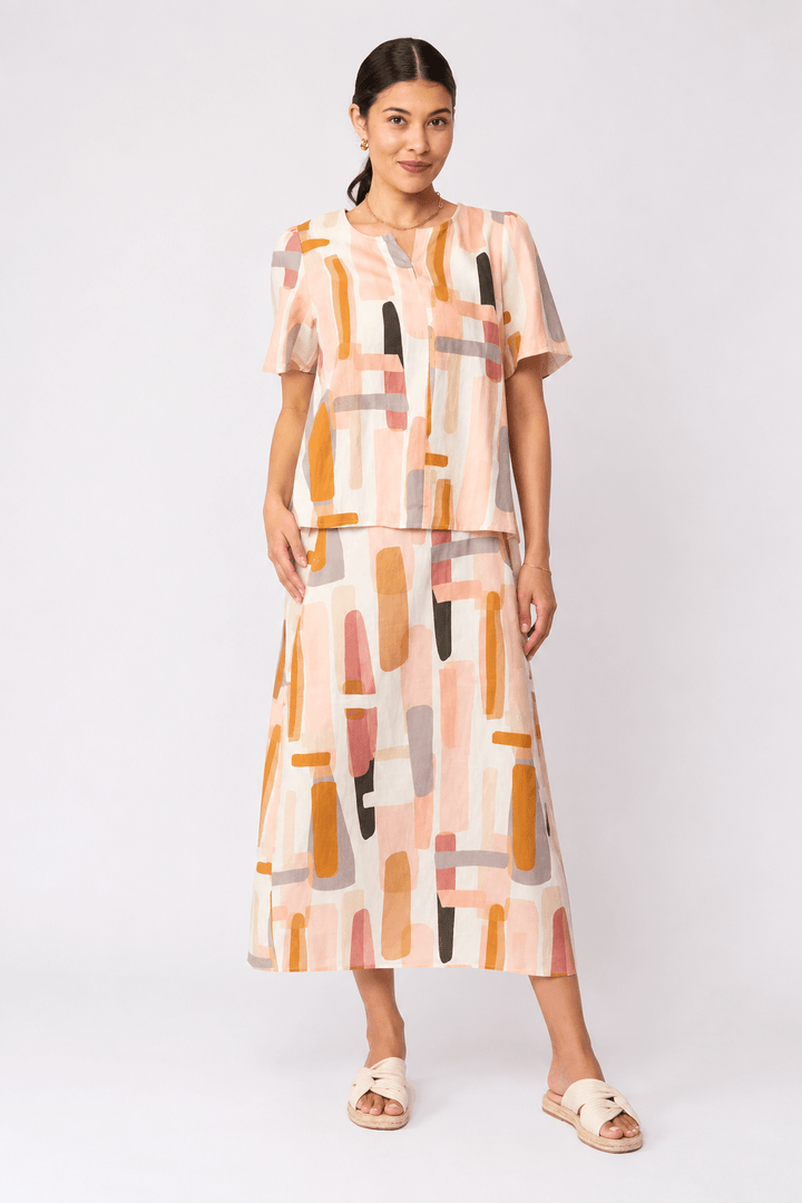 Foil model in a short-sleeve top with an abstract geometric print in peach, orange, beige, and gray, paired with matching skirt and black strappy sandals, against a light gray studio background.
Back view
