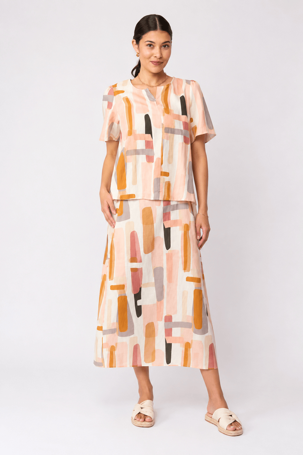 Foil model in a short-sleeve top with an abstract geometric print in peach, orange, beige, and gray, paired with matching skirt and black strappy sandals, against a light gray studio background.
Back view