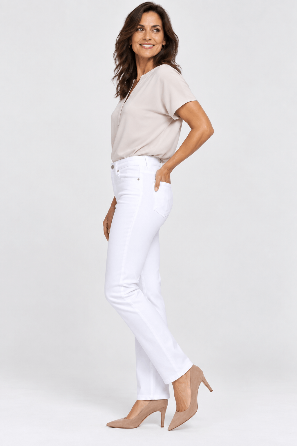 NYDJ model in a light beige short-sleeve henley top and white straight-leg jeans with beige heels, standing against a light gray studio background.