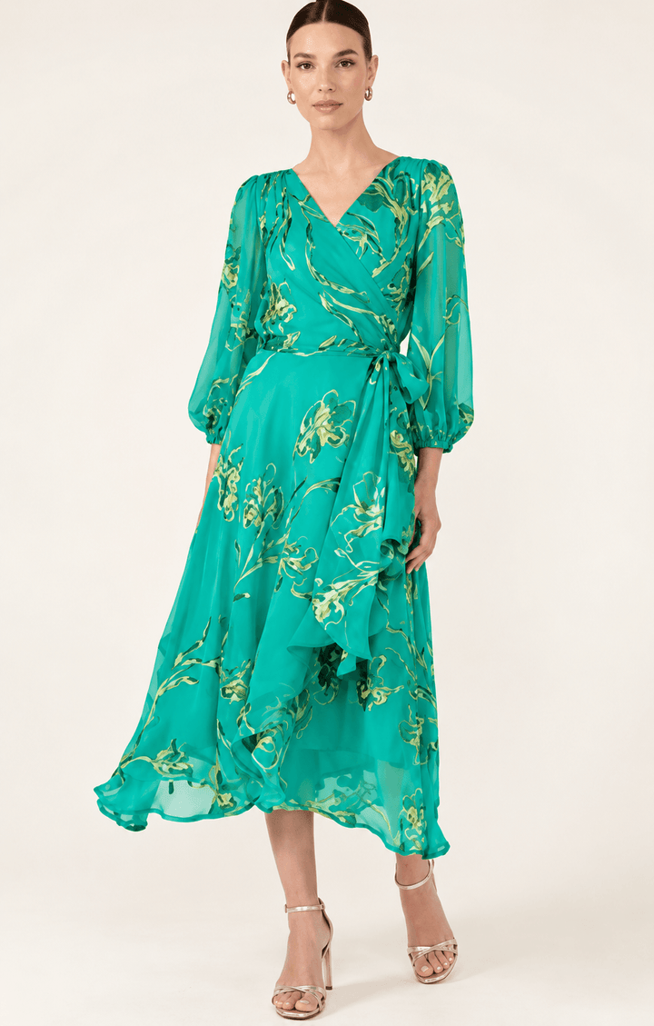 Sacha Drake modeling a flowing teal green midi dress with a gold floral print, three-quarter sheer sleeves, and a tie-waist detail, set against a plain light beige studio background.