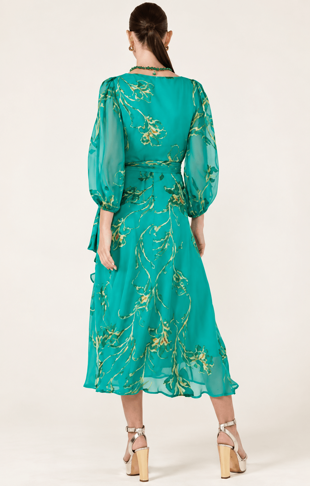 Sacha Drake modeling a flowing teal green midi dress with a gold floral print, three-quarter sheer sleeves, and a tie-waist detail, set against a plain light beige studio background. Back view