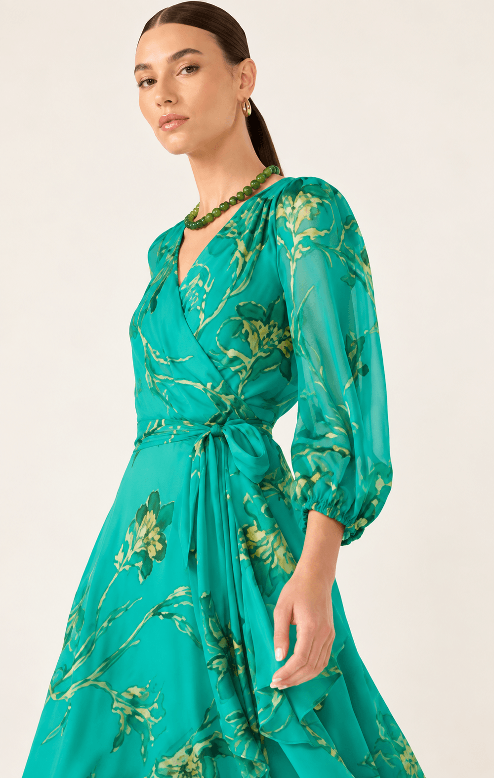 Sacha Drake modeling a flowing teal green midi dress with a gold floral print, three-quarter sheer sleeves, and a tie-waist detail, set against a plain light beige studio background.