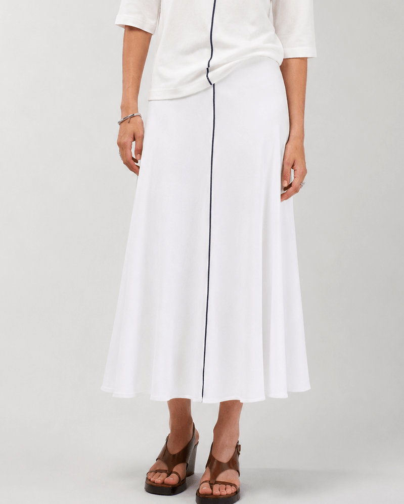 Madly Sweetly modeling a white two-piece outfit consisting of a short-sleeve top with dark navy trim and a matching midi skirt with a vertical navy stripe, paired with brown strappy sandals against a light gray studio background.