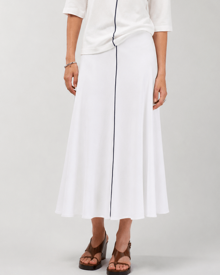 Madly Sweetly modeling a white two-piece outfit consisting of a short-sleeve top with dark navy trim and a matching midi skirt with a vertical navy stripe, paired with brown strappy sandals against a light gray studio background.