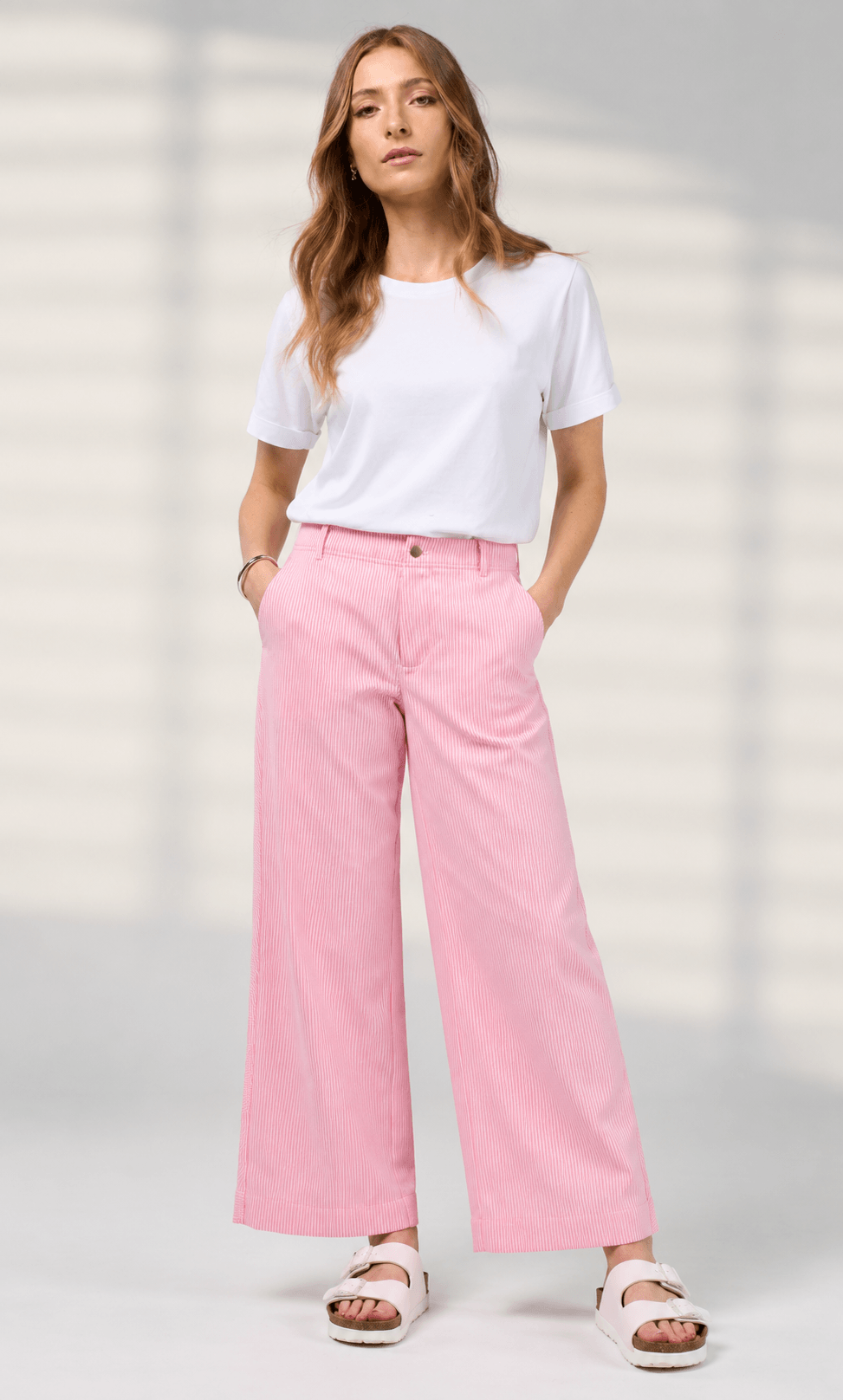 Madly Sweetly model wearing pink wide-leg pinstripe trousers, a white short-sleeve top, and white slide sandals, standing against a light neutral background.
