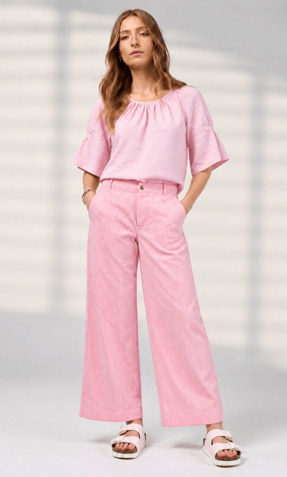 Madly Sweetly model wearing pink wide-leg pinstripe trousers, a white short-sleeve top, and white slide sandals, standing against a light neutral background.